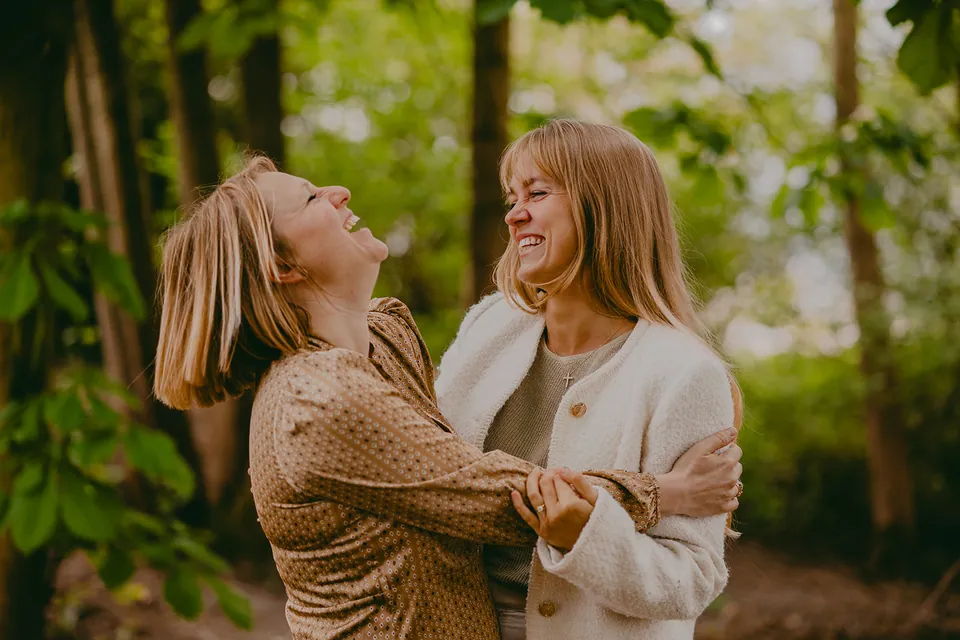 Twee vrouwen in camel en crème lachen samen tussen de bomen